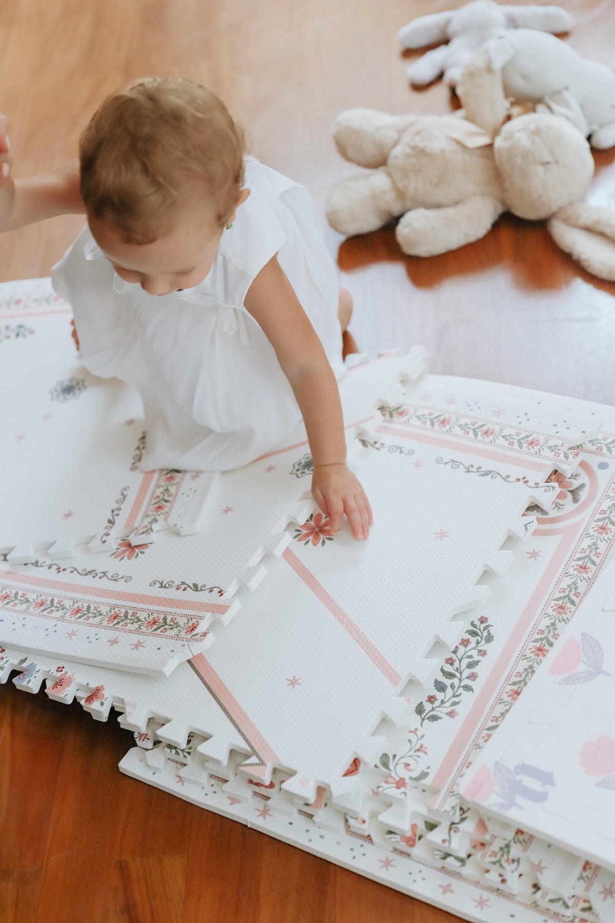 Bebê de vestido branco engatinhando sobre placas soltas do tapetinho Vintage Rose, com coelhos de pelúcia ao lado