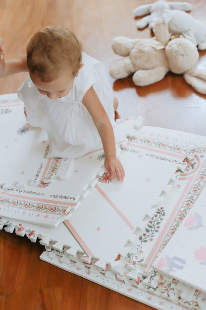 Bebê de vestido branco engatinhando sobre placas soltas do tapetinho Vintage Rose, com coelhos de pelúcia ao lado