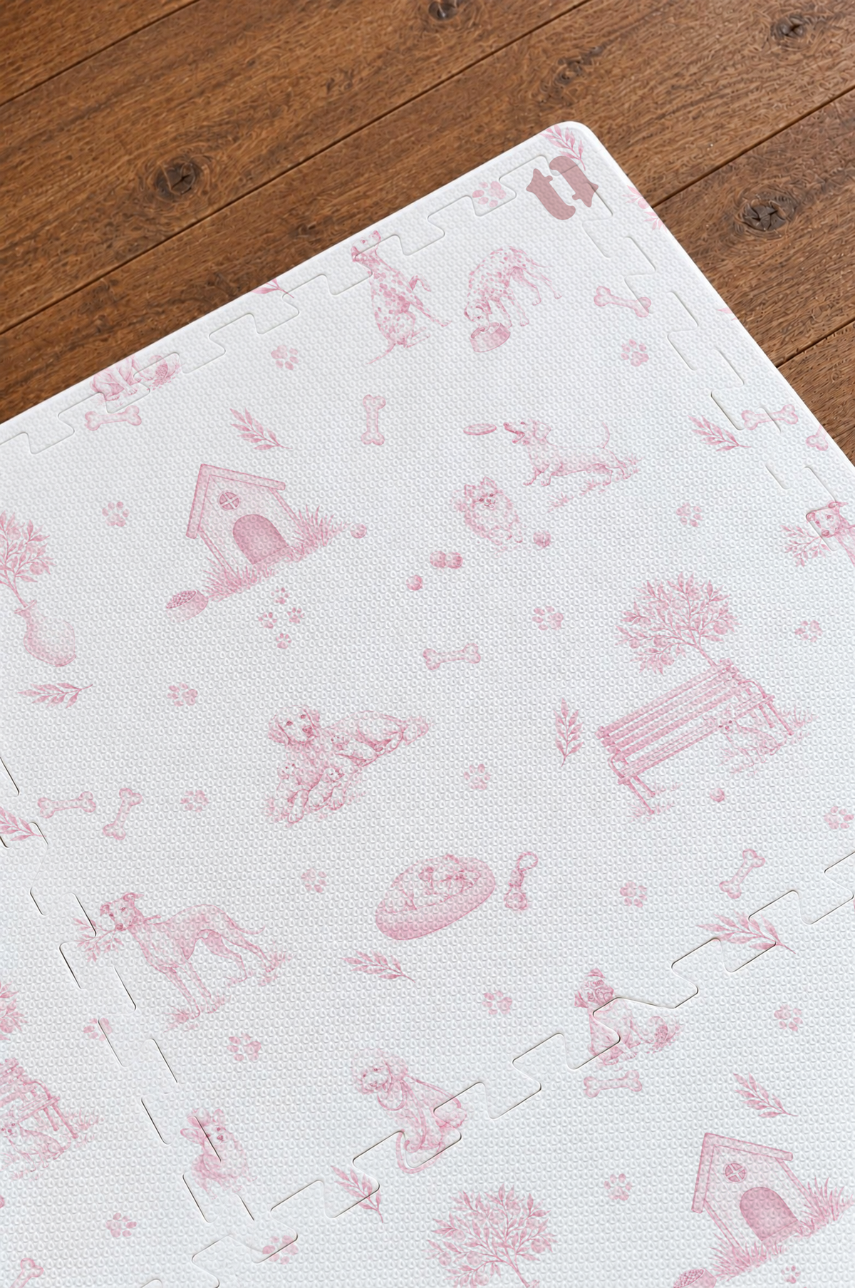 Close do canto do tapete infantil de EVA Toile de Jouy Dogs rosa montado sobre piso de madeira, com casinha, banco de praça, cachorrinhos, ossinhos e patinhas em rosa e os encaixes das placas