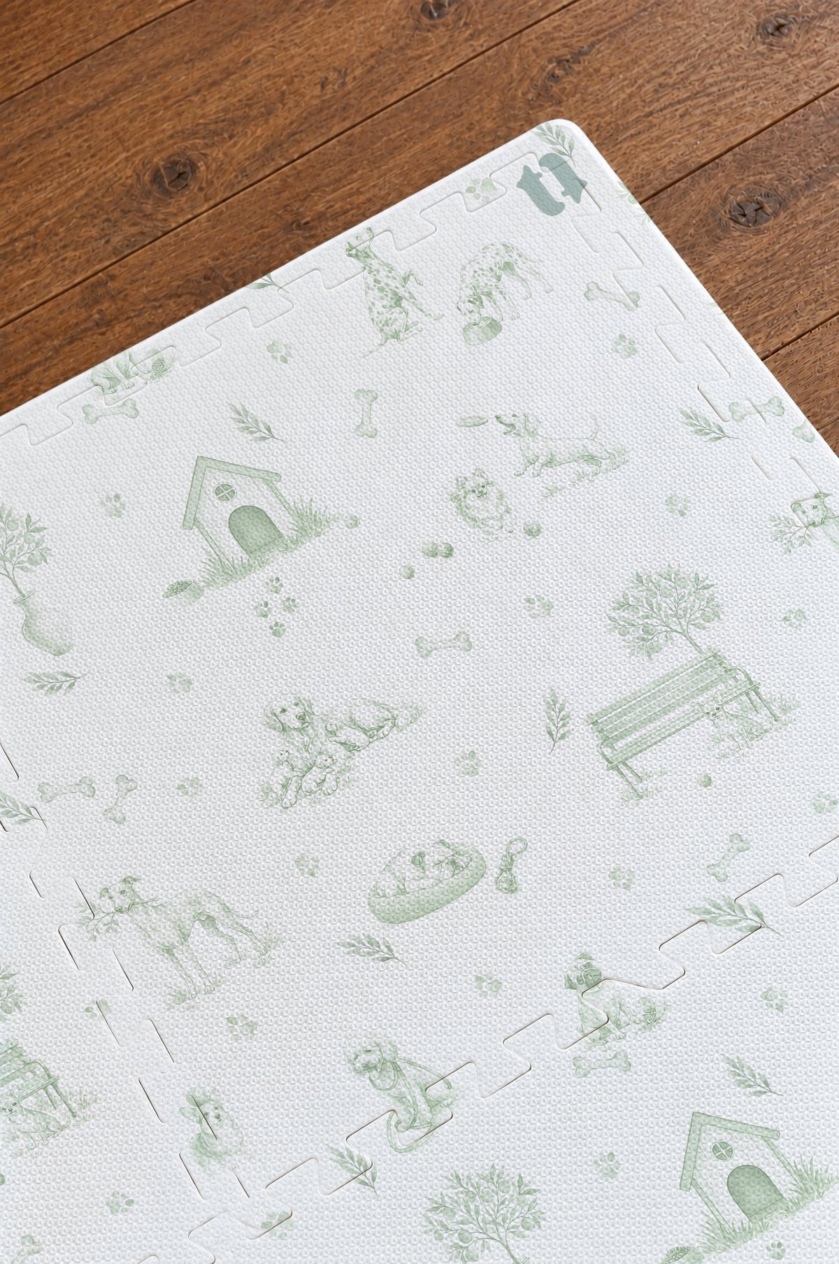Close do canto do tapete infantil de EVA Toile de Jouy Dogs verde montado sobre piso de madeira, com casinha, banco de praça, cachorrinhos, ossinhos e patinhas em verde e os encaixes das placas